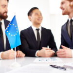 Image of European flag and business documents on workplace with three partners negotiating on background